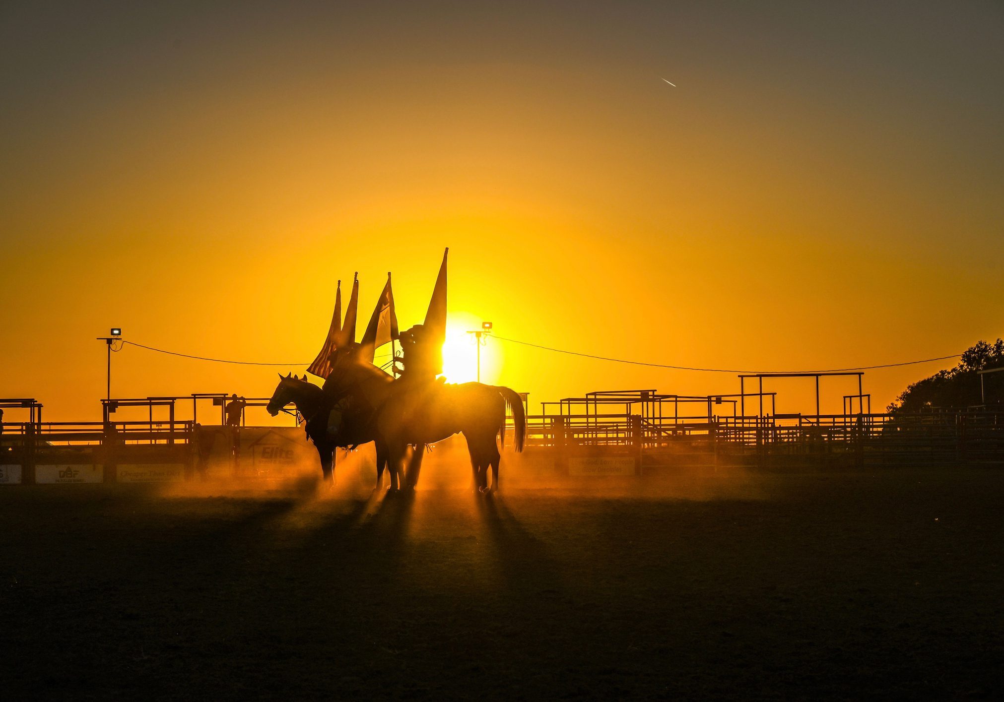 Silhouetted cowboys on horseback in a dusty rodeo arena at sunset with flags.